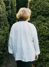 Load image into Gallery viewer, Older woman wearing French Cheesemaker's Smock made from white linen, standing by a large hedge with her back turned.