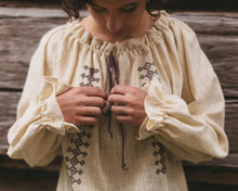 Load image into Gallery viewer, Picture of young woman adjusting ties of drawstring. Front view of Romanian blouse with gathered cuffs and neckline. Shirt is a natural gauze fabric with drawstring and embroidery applied in same color floss.
