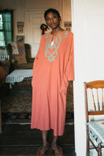 Load image into Gallery viewer, Young african american woman wearing long coral color version of 104 Egyptian shirt. Shirt has grey applique around neck and on front yoke of top. Model is leaning on door frame looking towards camera with hands in pockets.