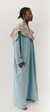 Load image into Gallery viewer, Black woman walking toward camera wearing a long blue dress with an embroidered triangular collar yoke.. She is standing sideways with a hand in a pocket and a white background.