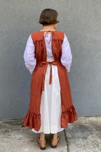 Load image into Gallery viewer, Back view of woman standing in front of a grey wall wearing a large sleeveless orange apron with pockets and a flounce at the bottom.