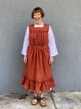 Load image into Gallery viewer, Woman standing in front of a grey wall wearing a large sleeveless orange apron with pockets and a flounce at the bottom.