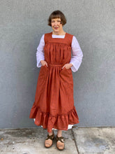 Load image into Gallery viewer, Woman standing in front of a grey wall wearing a large sleeveless orange apron with pockets and a flounce at the bottom.
