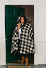 Load image into Gallery viewer, Black woman standing in a door wearing a large black and white checked burnoose with the hood up.