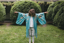 Load image into Gallery viewer, Child wearing a blue floral kimono standing in a garden with green bushes.
