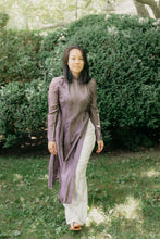 Load image into Gallery viewer, Vietnamese American woman wearing a purple silk Ao Dai tunic with white pants and sandals. She is walking toward the camera in a lawn with shrubs in the background.