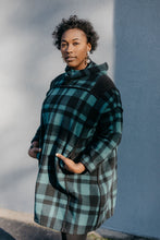 Load image into Gallery viewer, Black woman standing outside in front of a grey wall wearing a green and black plaid siberian parka. Her hands are in the pockets.