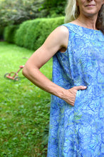 Load image into Gallery viewer, White woman wearing a sleeveless blue batiked muumuu, standing outside by shrubbery. her hand is in her pocket and view is close up on the pocket.