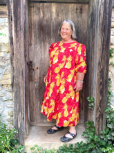 Load image into Gallery viewer, Older woman wearing a red muumuu with yellow pineapples on it. Sleeves are 3/4 length and she is standing in a wooden doorway.