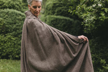 Load image into Gallery viewer, Older white woman with gray hair standing surrounded by greenery wearing 207 Kinsale Cloak without hood holding the cloak out with one arm to show length.