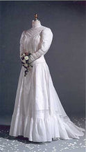 Load image into Gallery viewer, Mannequin wearing 227 Edwardian Bridal Gown and Dress standing in front of a gray studio backdrop, holding a bouquet, with white flower petals sprinkled on the floor.