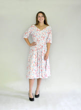 Load image into Gallery viewer, Young woman smiling wearing a below-knee length white and pink floral print dress with a v-neck and 3/4 length sleeves and black shoes. She is in front of a white background.