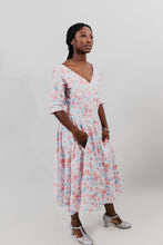 Load image into Gallery viewer, Young woman wearing a below-knee length white and pink floral print dress with a v-neck and 3/4 length sleeves and black shoes. She is in front of a white background and her hands are in the skirt pockets.