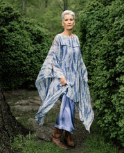 Load image into Gallery viewer, Older white woman with short grey hair standing surrounded by greenery,wearing brown boots and the Greek Island Dress Tunic displaying the flare of the free- floating sleeve panels.