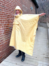 Load image into Gallery viewer, Woman standing a yellow rain poncho by a brick wall with her arm to the side to show how wide the poncho is.