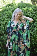 Load image into Gallery viewer, White woman wearing a tropical green and orange print kaftan standing out side in front of greenery with her hand on her neck.