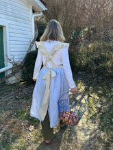 Load image into Gallery viewer, Outside in greenery the back of a woman in a white long sleeved shirt and green pants wearing the light blue linen apron with beige ruffled straps , pockets and waistband holding a basket of eggs in her right hand.