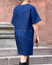 Load image into Gallery viewer, Person wearing a blue denim dress standing on stone steps with a red-brown wall in the background.