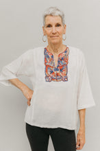 Load image into Gallery viewer, Older woman in short version of Egyptian top.  Top is made in a white gauze fabric with multi-colored embroidery on yoke.  Model has one hand on hip and poses with white backdrop.
