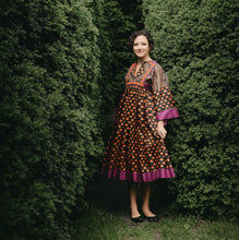 Load image into Gallery viewer, Young woman standing between two bushed with floral poppy print Afghan Nomad dress.
