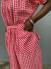Load image into Gallery viewer, Close up of hand in a pocket of a woman standing in front of grey wall wearing a red and white check dress with short puffed sleeves and a bottom flounce.
