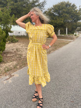 Load image into Gallery viewer, Woman wearing a yellow and white checked calf-length dress with puffed sleeves and pockets.  She is standing in a street with a hand by her head.  Dress has a flounce at the bottom
