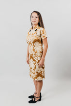 Load image into Gallery viewer, Asian woman wearing a gold brocade knee-length cheogsam, standing sideways in front of a white background.

