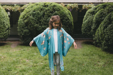 Load image into Gallery viewer, Young girl wearing a blue floral kimono standing in a garden with manicured hedges.
