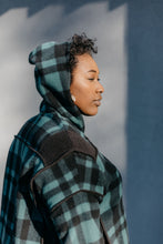 Load image into Gallery viewer, Close up of Black woman standing outside in front of a grey wall wearing a green and black plaid siberian parka. The hood is up and she is facing to the right.
