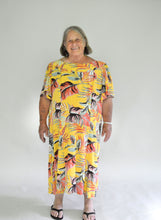 Load image into Gallery viewer, Older woman wearing a size 2XL knee-length, short-sleeve, yellow muumuu with orange and green palm fronds on it.

