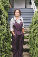 Load image into Gallery viewer, Young brunette white woman standing with hands in pockets of the 240 Rosie the Riveter overalls surrounded by greenery.
