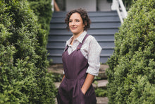 Load image into Gallery viewer, Young brunette woman turned slightly to the left wearing the Rosie the Riveter overalls with hands in the pockets surrounded by greenery.
