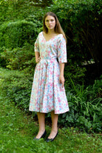 Load image into Gallery viewer, Young woman standing in front of trees and shrubs wearing a white and pink floral print dress with a v-neck.
