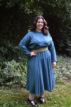 Load image into Gallery viewer, Young woman wearing a blue long sleeve, jewel neck, calf-length dress outside in front of shrubbery.  She has on a green a white beaded belt and black shoes. Her right hand is on her hip.
