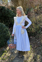 Load image into Gallery viewer, A woman standing outside with greenery in the background. She is wearing the  blue linen apron with beige ruffled straps , pockets and waistband. She has her left hand on her hip and a basket of eggs being held with her right hand
