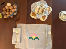 Load image into Gallery viewer, On a wooden table is a basket of eggs on the left top corner, biscuits in a biscuit cozy on the center top, a silver candle stick on the right top corner. On the center bottom there is a napkin with a silver fork on top, a placemat with a painted flower in pink yellow and green with a knife and spoon on top.
