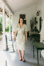 Load image into Gallery viewer, Woman walking towards the camera out doors on a sun lit porch.  There are tables in the foreground and chairs and a table in the background.  She is wearing a mint green and lavender floral dress.  Dress is View B. View B is more casual, with buttoned front, flounce collar and sleeves, and eyelash pockets.
