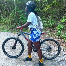 Load image into Gallery viewer, Person wearing blue floral biking shorts with a bicycle on a forest path
