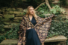 Load image into Gallery viewer, Young white blonde woman sitting on a stone bench with left hand stretched on a stone wall wearing the Poiret Cocoon Coat
