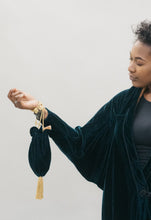 Load image into Gallery viewer, An African America woman wearing a velvet dark green Poiret Cocoon Coat against a studio white backdrop, holding a dark green velvet drawstring bag with gold cording.
