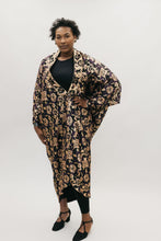 Load image into Gallery viewer, African American woman standing with left hand on her hipin front of a studio white backdrop wearing the 503 Poiret Cocoon Coat in a silk charmeuse fabric with gold roses as the pattern. She is also wearing black closed toed shoes.

