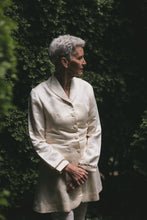 Load image into Gallery viewer, Older white woman with grey hair standing with hands in the front one on top of the other,looking to her left surrounded by greenery wearing the Countryside Frock Coat buttoned in white.
