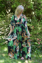 Load image into Gallery viewer, White woman wearing a tropical green and orange print kaftan standing out side in front of greenery and white and pink flowers.
