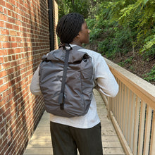 Load image into Gallery viewer, Person wearing a gray backpack on a wooden deck with a brick wall and greenery in the background
