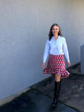 Load image into Gallery viewer, Woman standing in front of a grey wall outside, wearing a buttondown shirt with the Flamenco practice skirt and black boots.  Skirt is red and white ankara fabric and is above the knee in the front and top of calf in the back with a ruffled flounce.  Woman is walking.

