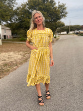 Load image into Gallery viewer, Woman wearing a yellow and white checked calf-length dress with puffed sleeves and pockets.  She is standing in a street and has a hand in her Woman wearing a yellow and white checked calf-length dress with puffed sleeves and pocket.
