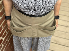 Load image into Gallery viewer, Person wearing a beige apron tool belt with multiple pockets against a brick wall.
