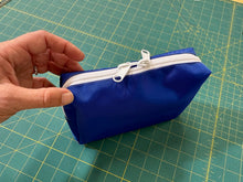 Load image into Gallery viewer, Blue zippered pouch with white zipper held by a hand on a green cutting mat.

