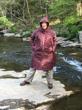 Load image into Gallery viewer, Woman standing on a rock in a creek wearing the parka made of maroon waxed cotton . Her hands are in her pockets.
