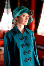 Load image into Gallery viewer, Close up of a young blonde woman in a red lounge area., wearing the Metropolitan suit with front closing frogs, and a hat.
